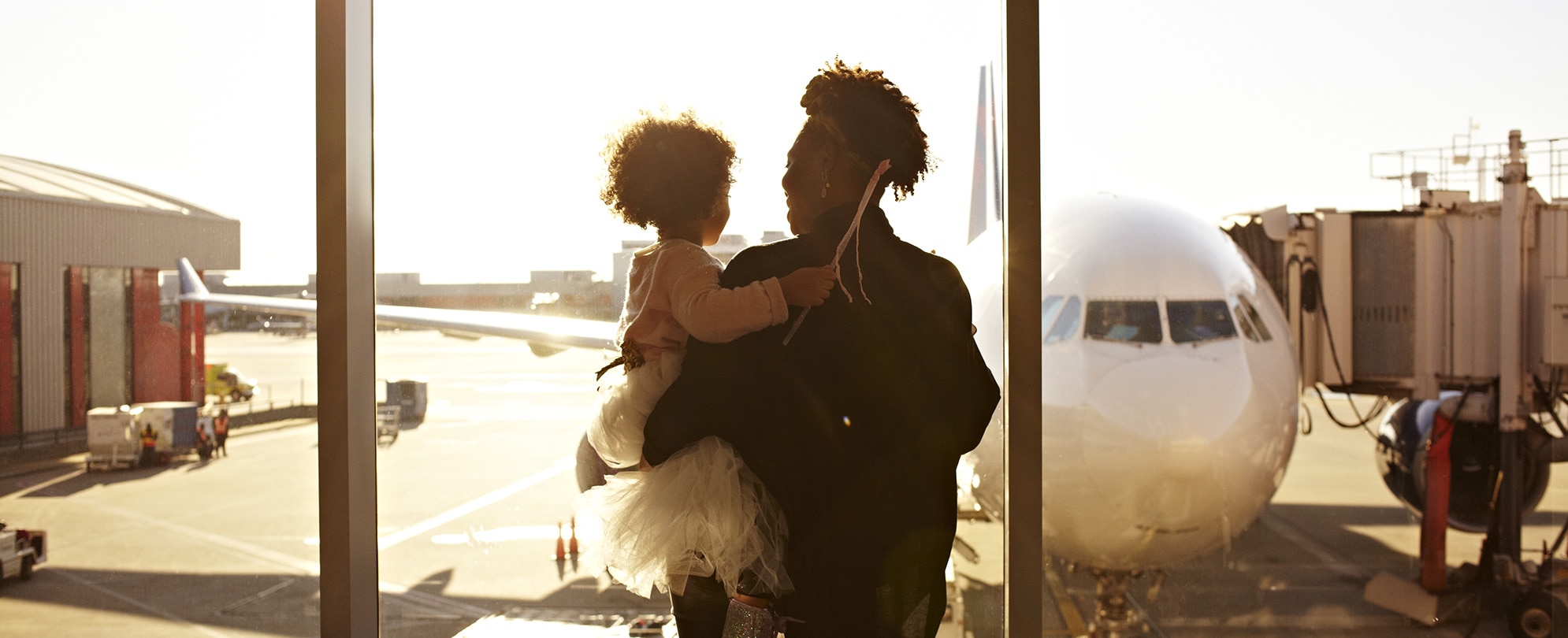 A mom holds her young daughter while looking out through the Atlanta Airport windows at an airplane and jet bridge.